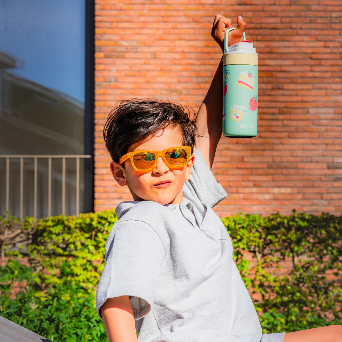 Enfant qui brandit à bout de bras sa gourde Lagoon Insulated 400 ml Picnic Party devant un mur de briques au soleil